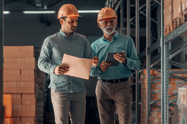 Two men in hardhats and blue shirts in a warehouse with a folder and tablet.