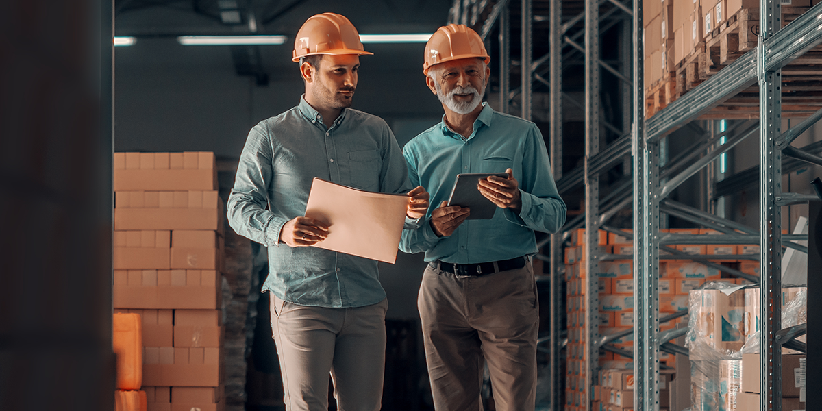 Two men in hardhats and blue shirts in a warehouse with a folder and tablet.