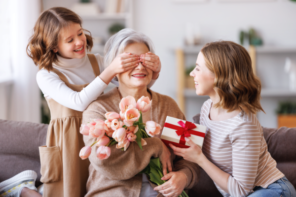 A little girl covers her grandmother's eyes as she is holding flowers while a third woman presents her a wrapped gift