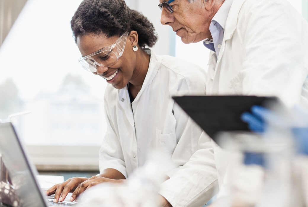 Woman in lab coat and safety goggles typing on computer with man overseeing