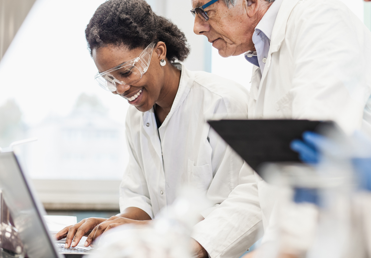 Woman in lab coat and safety goggles typing on computer with man overseeing