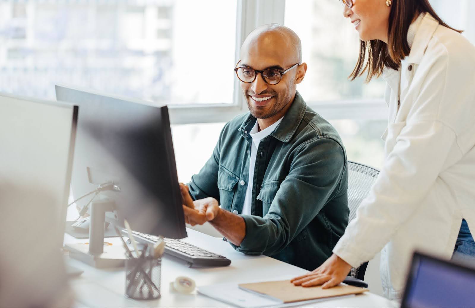 Man sitting at desk pointing at computer monitor and smiling with woman standing next to him leaning one hand on the table