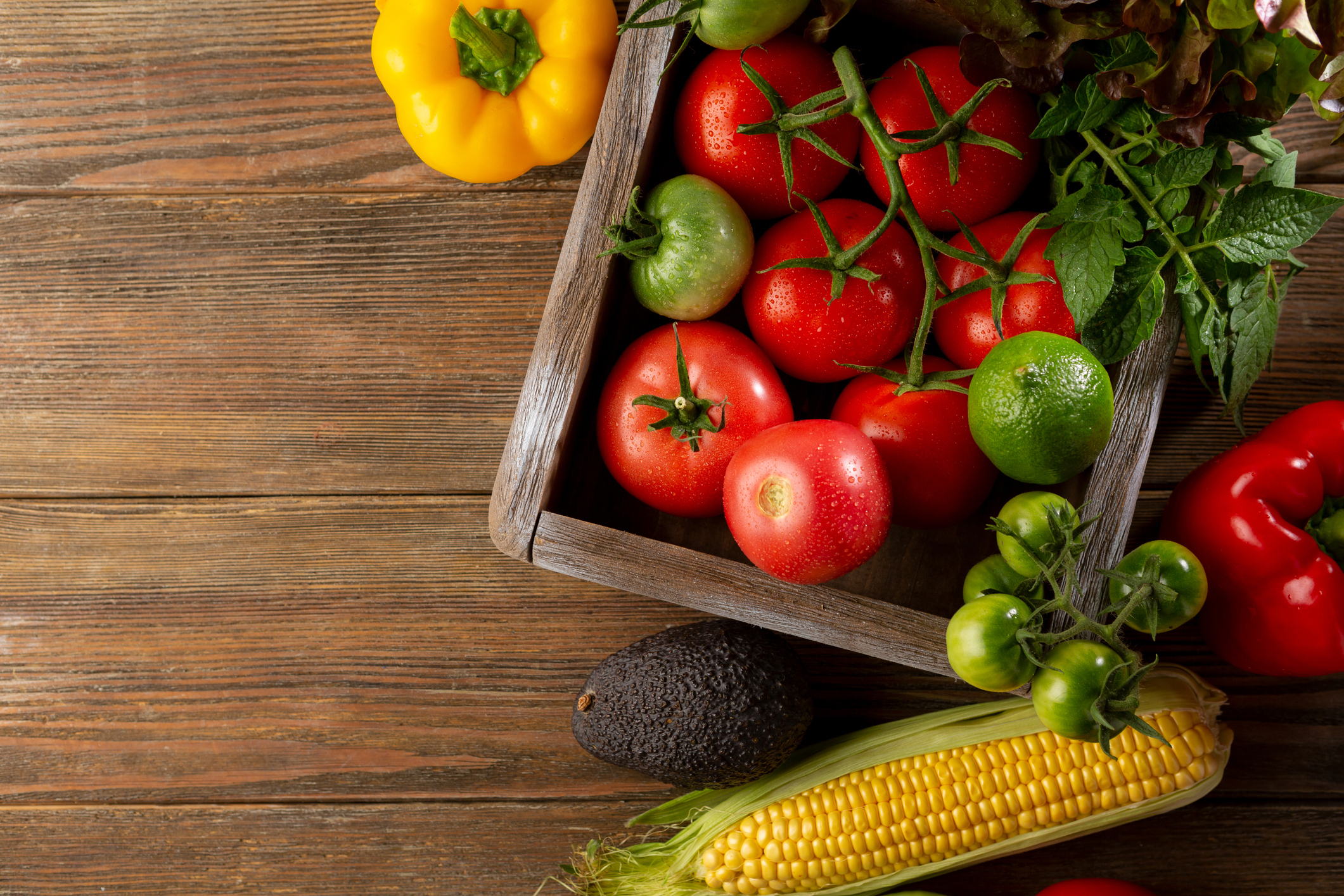 A box with tomatoes, lime, mint on a table with an avocado, corn and bell peppers next to it
